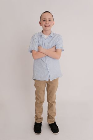 Smiling boy standing with arms crossed wearing a blue striped shirt and khaki pants against a white background.