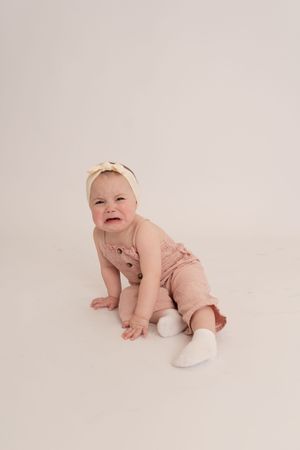Crying baby girl in pink romper and bow headband sitting on white studio floor.
