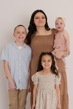 A young mother in a brown dress poses with three children, including a baby girl, against a neutral background.