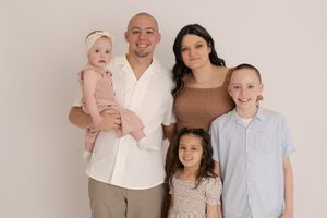 Family of five posing together in a bright studio, parents with three young children smiling warmly in neutral tones.
