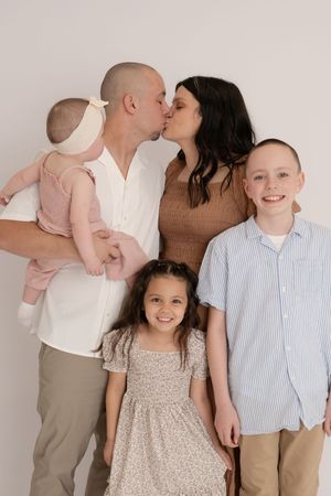 Family portrait with parents kissing while holding baby and two children smiling.