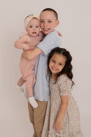 Three siblings posing together, older boy holding baby girl with younger sister beside them.