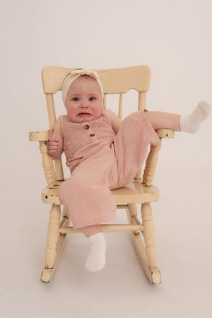 Adorable baby girl in a pink outfit and white headband sits in a small wooden rocking chair.
