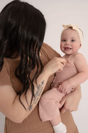 Dark-haired tattooed mother holding a smiling baby girl in a pink romper with a cream bow headband.