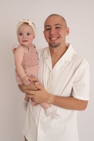 Smiling bald man holding a baby girl in pink outfit with bow headband against light background.