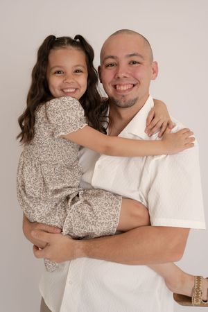 Smiling bald man holding a happy young girl in a floral dress, sharing a warm embrace in a studio photo.