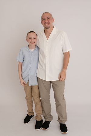 Smiling man in white shirt stands with young boy in blue shirt against white background.