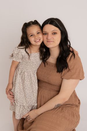 Dark-haired woman in brown dress poses with smiling young girl in floral dress.