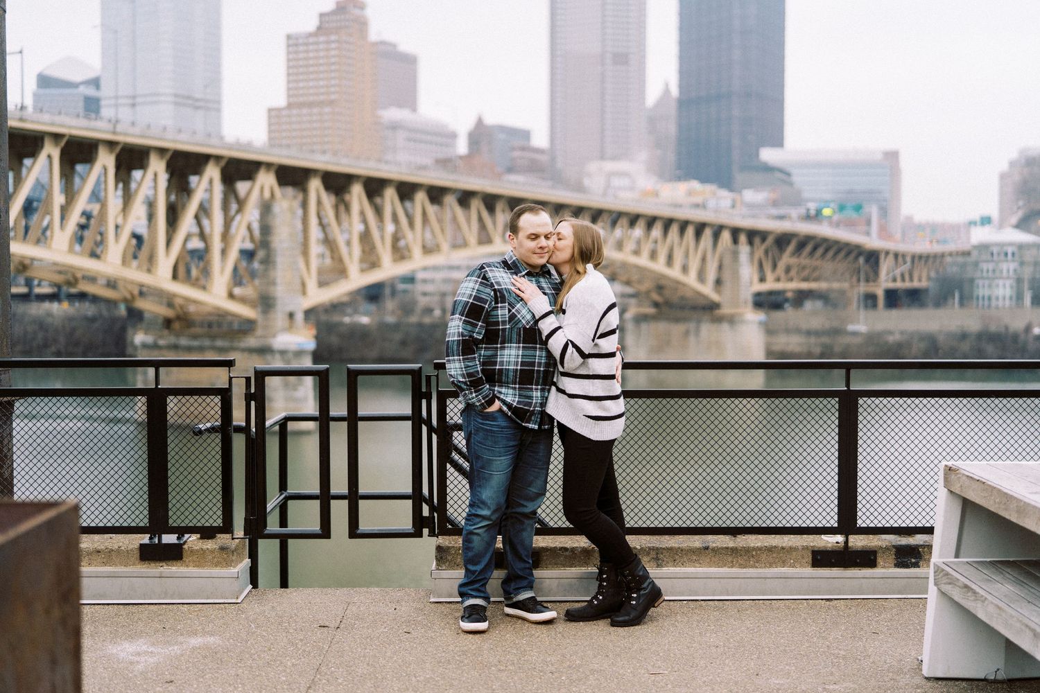 Couple embracing on a waterfront deck with a bridge and city skyline in the background on a cloudy day.