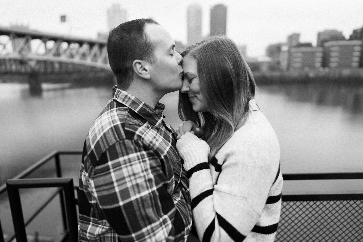 Romantic couple nearly kissing on a bridge with a city skyline and river in background.