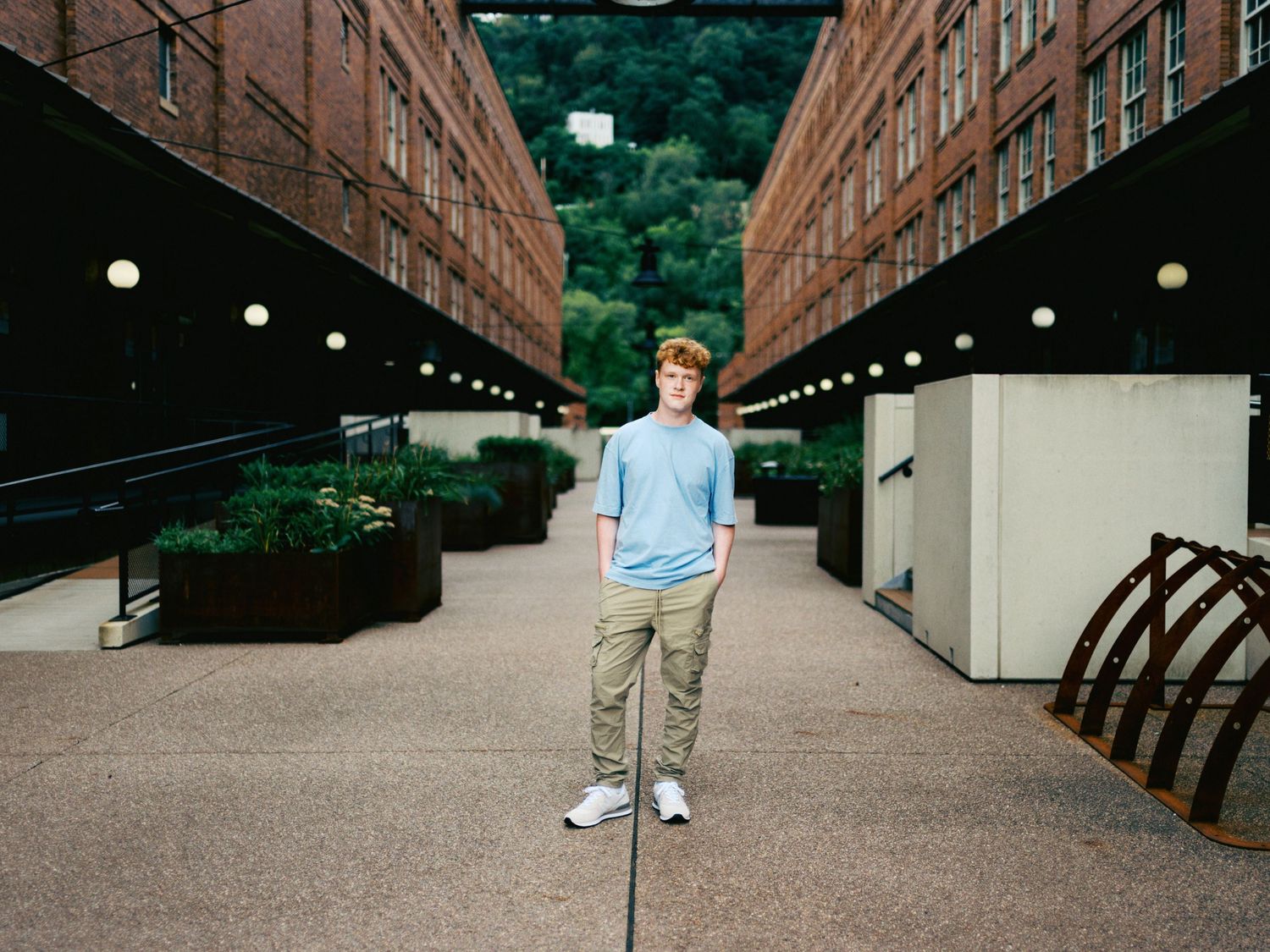 Young man with red hair in light blue shirt and khaki pants stands between two brick buildings.