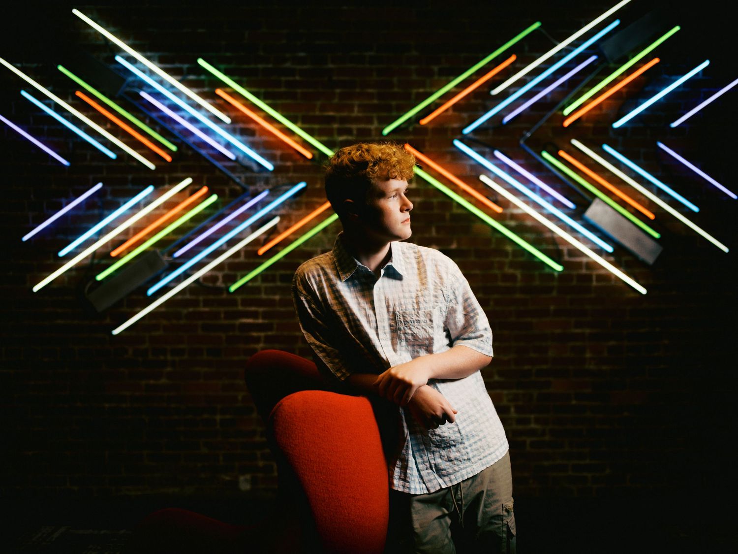 Young man posing in front of colorful neon light art installation on brick wall.
