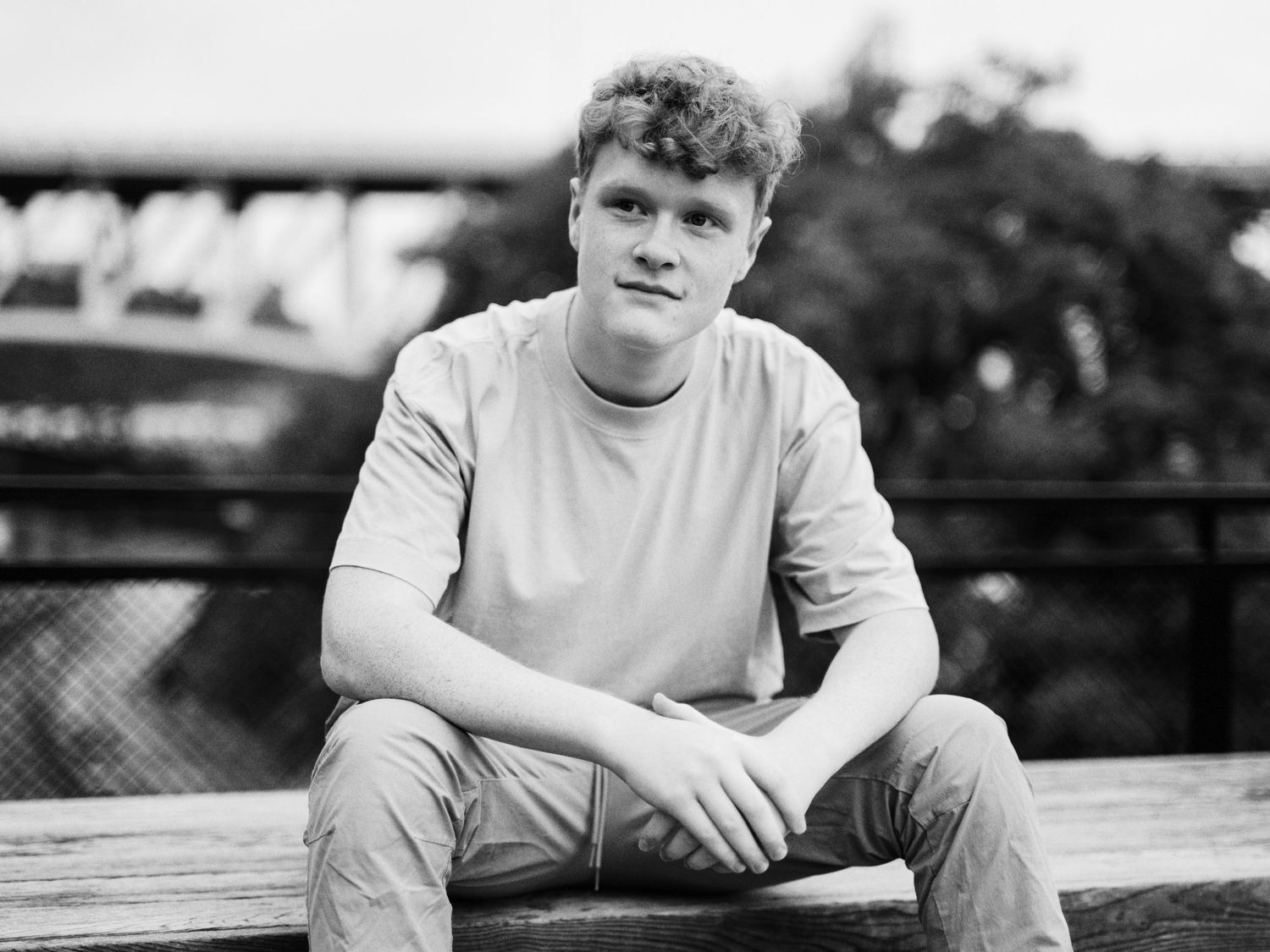 Young man in casual t-shirt sitting relaxed on wooden bench outdoors in black and white portrait photo.