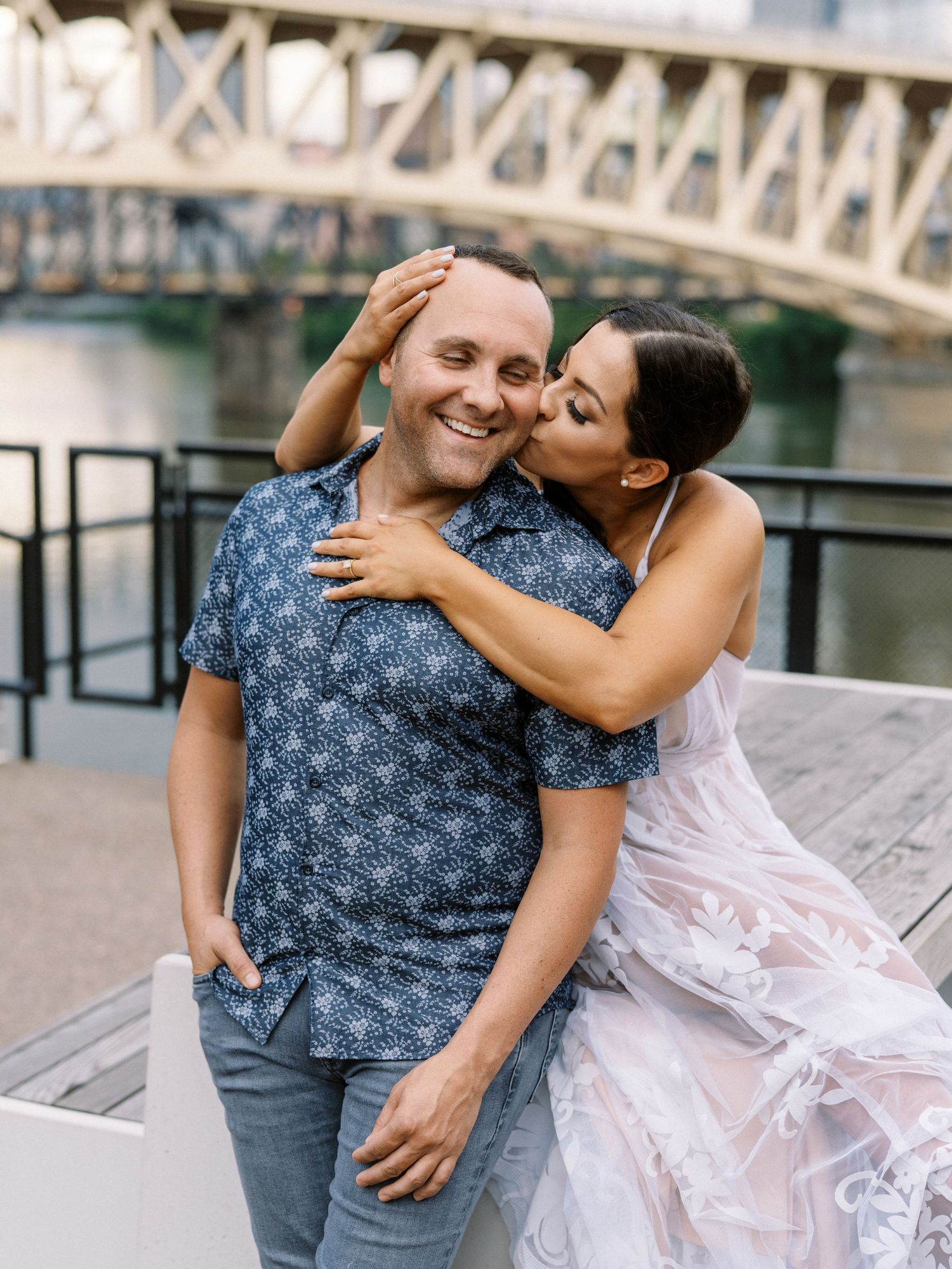 A couple shares a romantic moment by a river bridge, woman in pink dress kissing mans cheek as he smiles warmly.