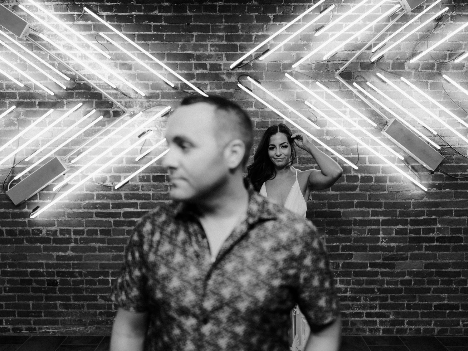 Black and white photo of a man in floral shirt and woman posing in front of neon arrow signs on brick wall.