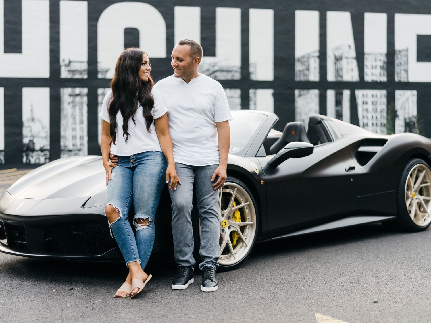 Couple posing beside a black Ferrari 488 Spider sports car in front of a urban mural backdrop.
