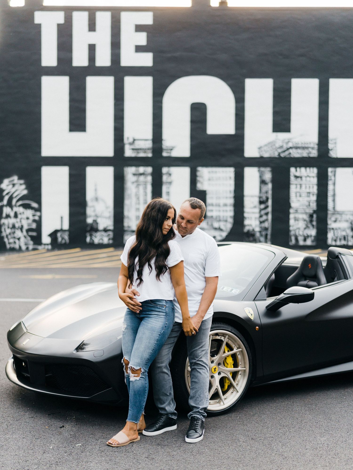 Couple posing in front of a black Ferrari sports car with a bold street mural backdrop in an urban setting.