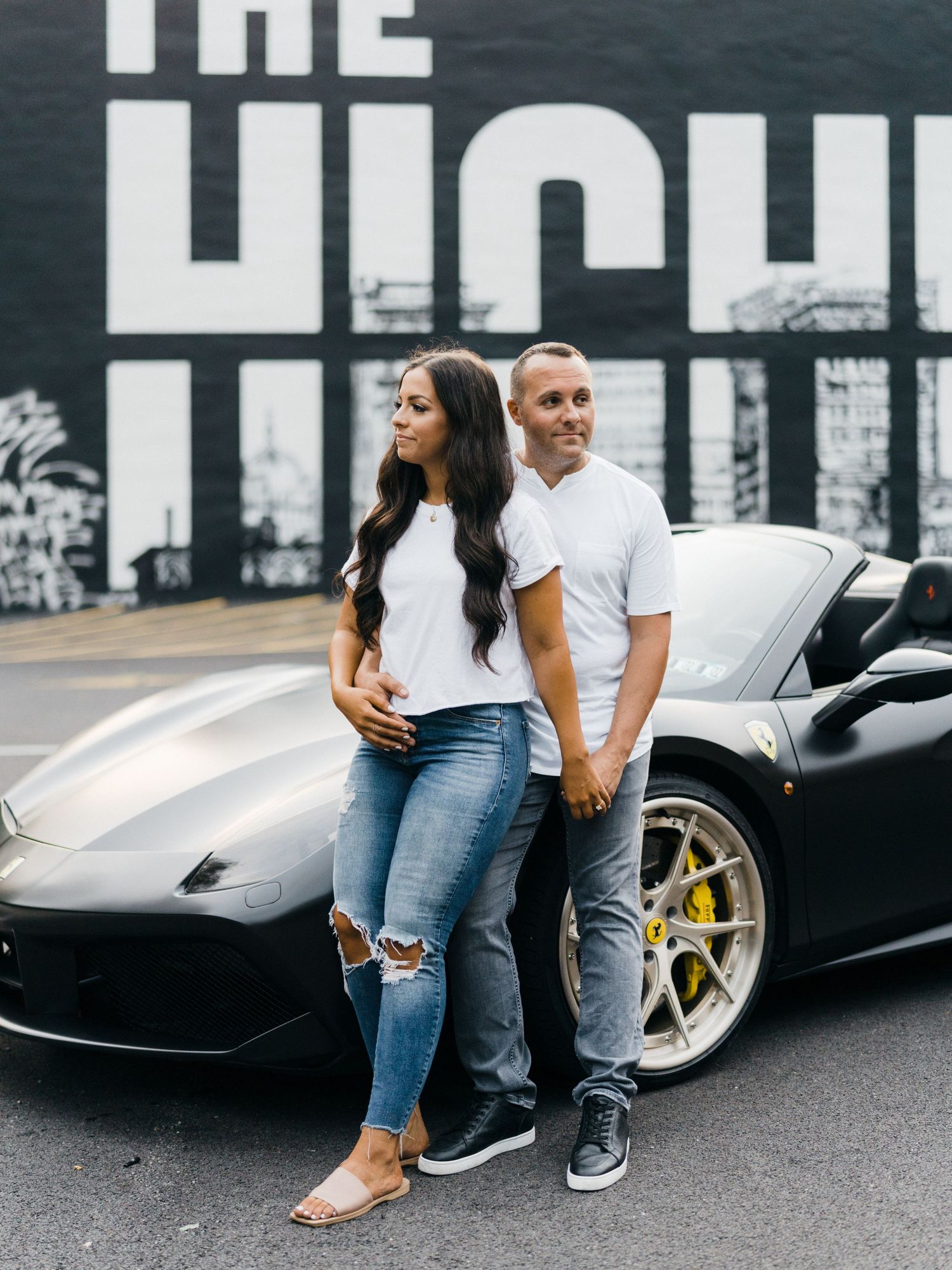 Stylish couple in matching white tees posing confidently in front of a sleek black Ferrari sports car.