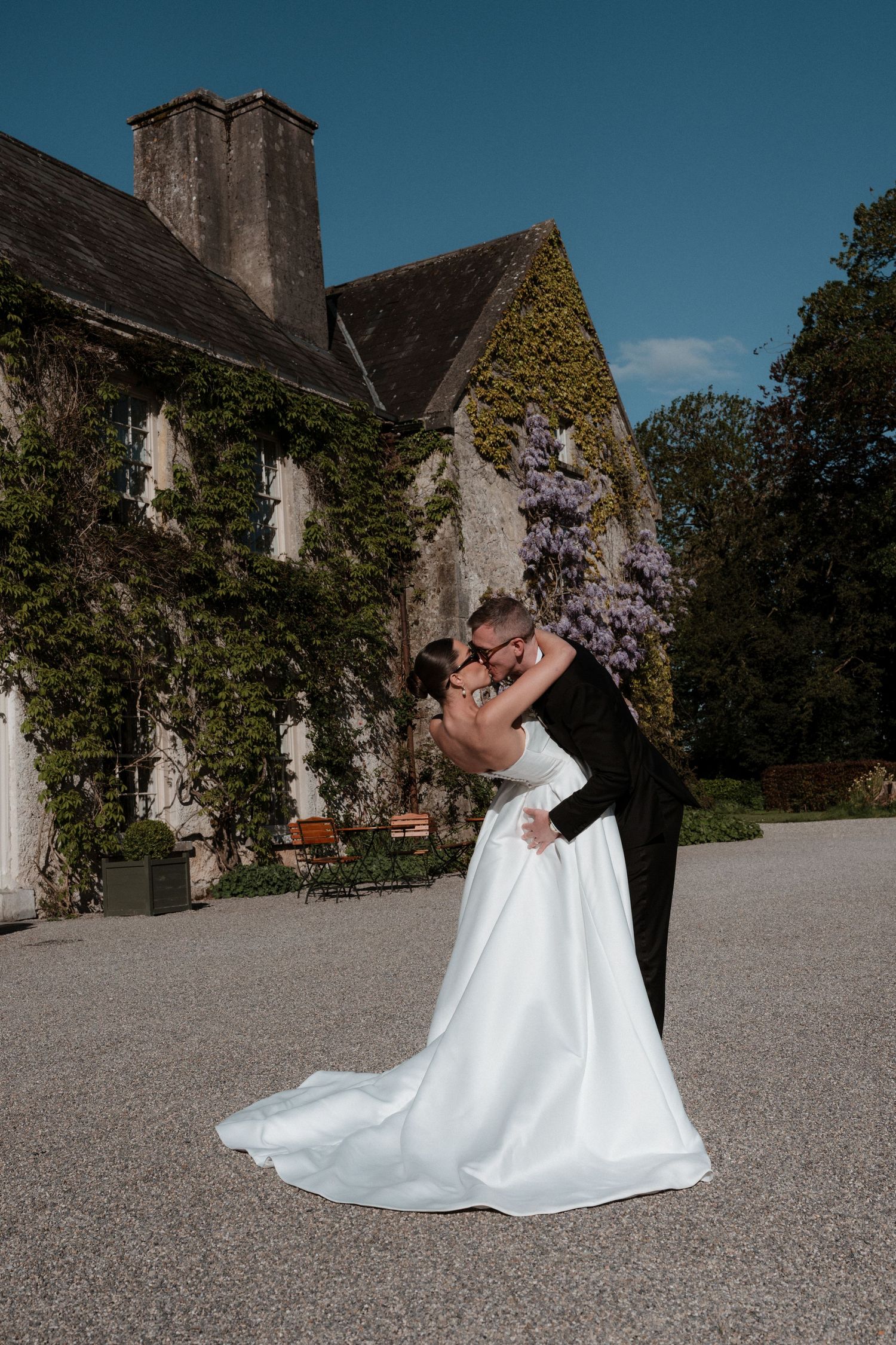 Bride and groom share a romantic embrace outside an ivy-covered stone manor under a bright blue sky.