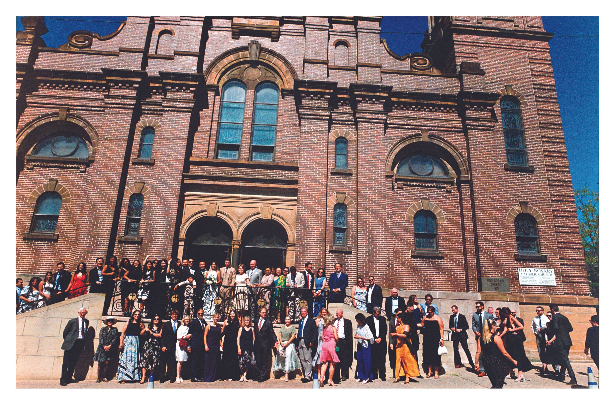Large group photo of formally dressed people on steps of grand brick church exterior.