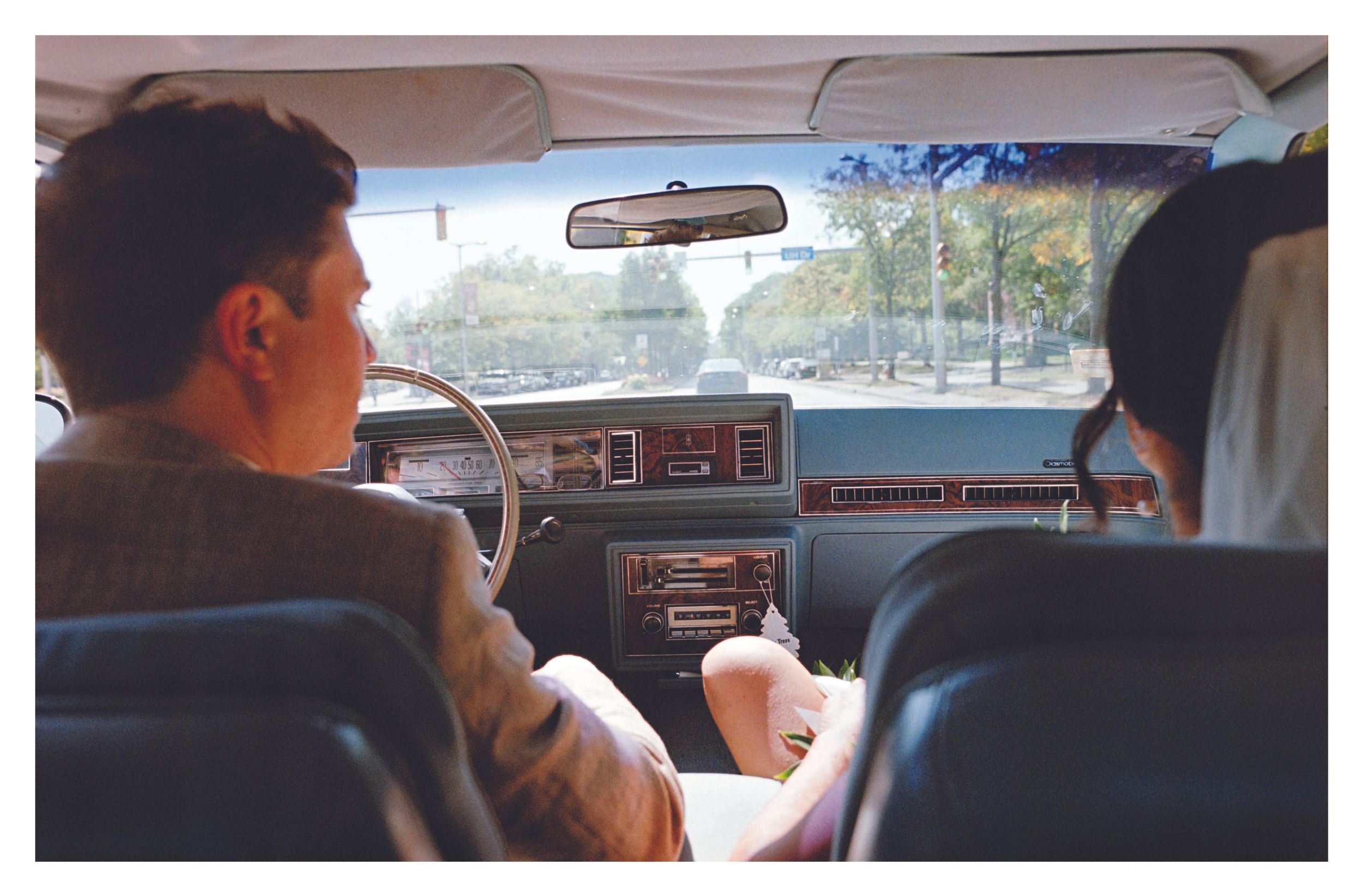A couple in wedding attire rides together in the front seat of a vintage car.