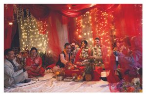 A traditional South Asian wedding ceremony with the couple seated beneath red drapes and fairy lights.