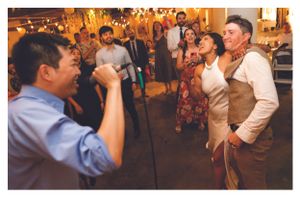 Guests laugh and sing together at a lively indoor wedding reception with warm string lights.