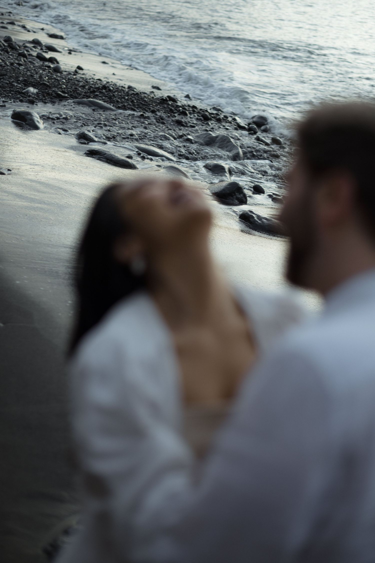 Romantic couple in white embracing on a rocky beach at dusk, woman tilting head back joyfully.