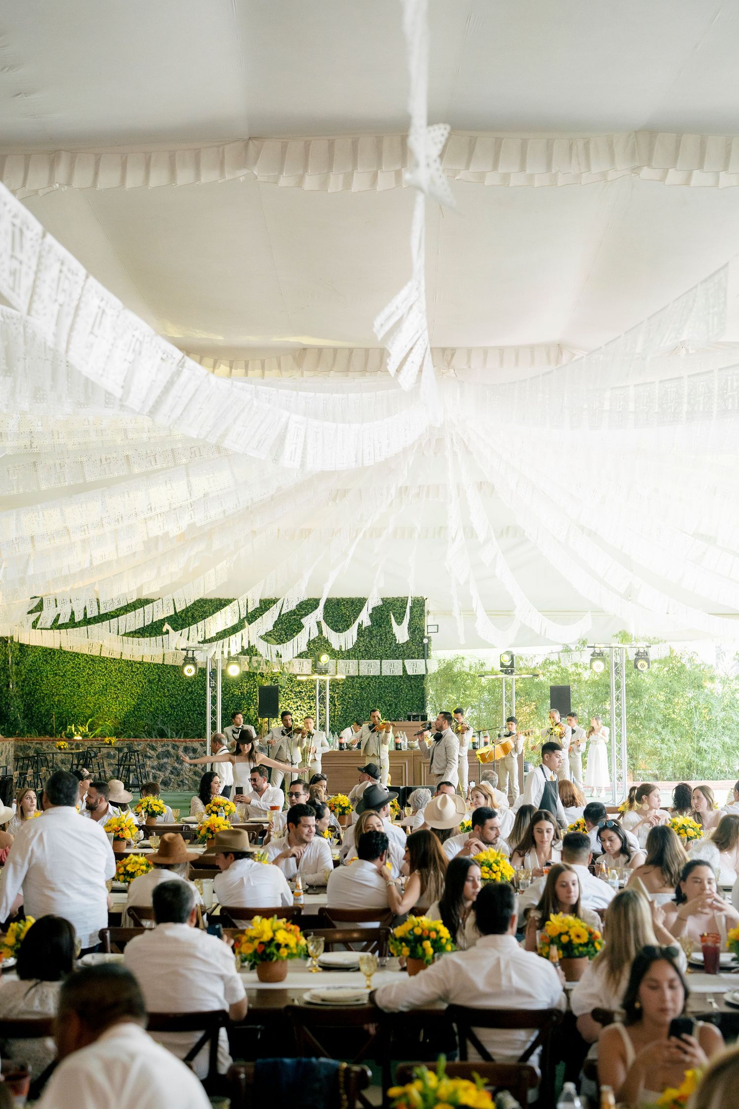 Elegant outdoor wedding reception under white draped tent with yellow floral centerpieces and guests dressed in white attire.