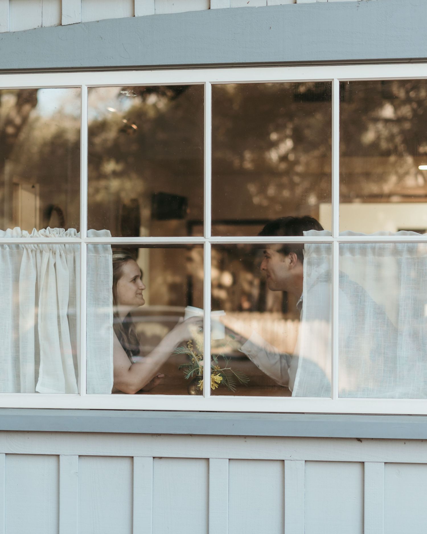 Couple sharing an intimate moment seen through a white-framed window with sheer curtains and warm natural light.