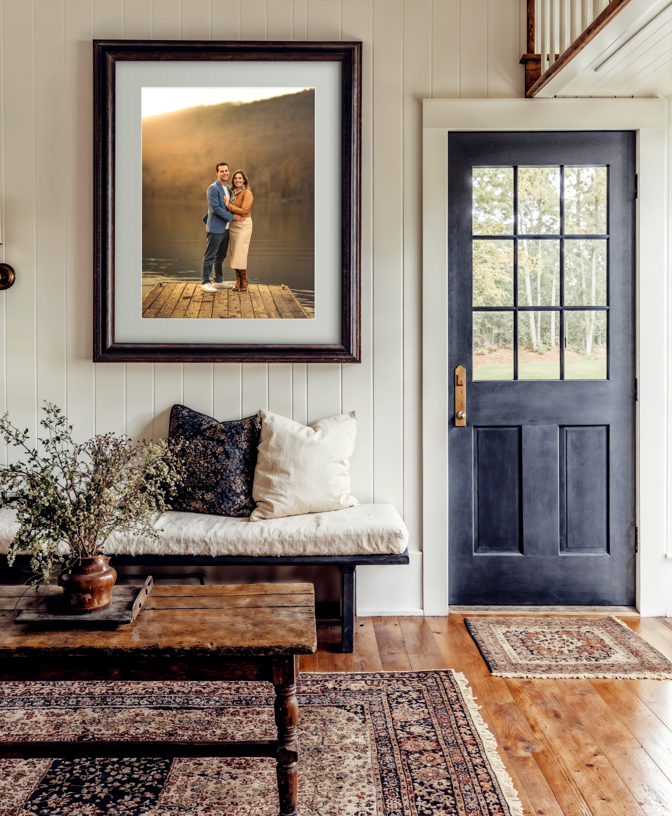 Cozy farmhouse entryway with framed couple portrait, navy door, wooden bench, Persian rug, and rustic coffee table.