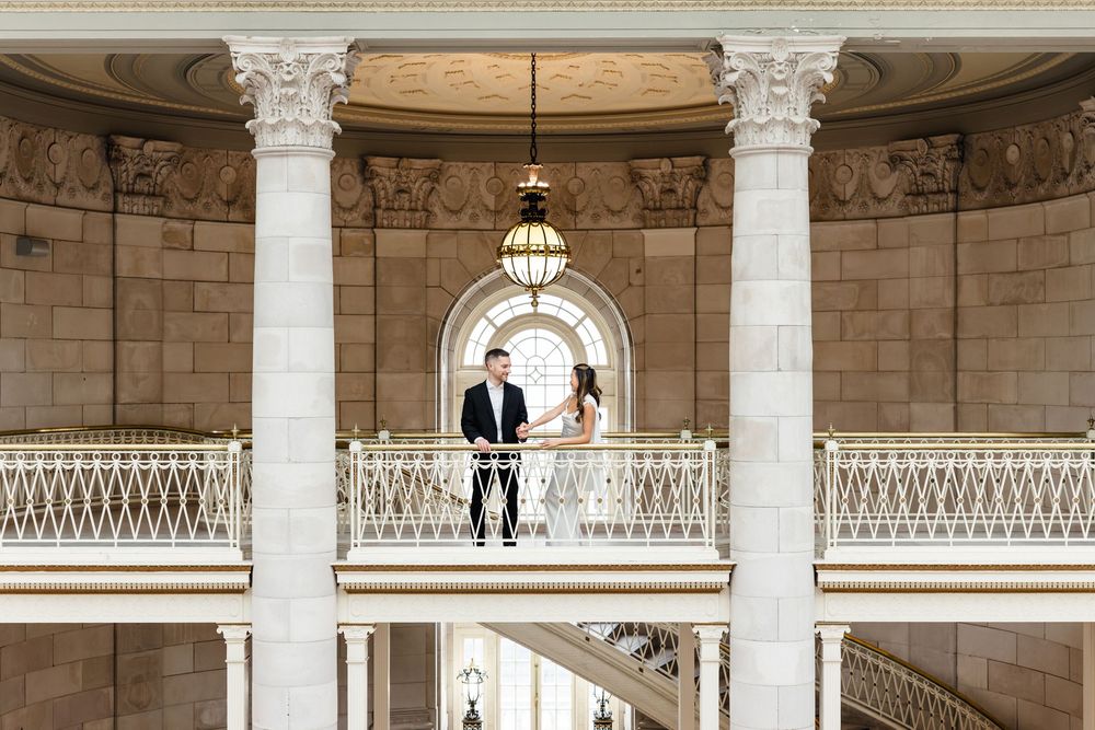 Couple standing on ornate balcony with white columns in elegant neoclassical building interior.