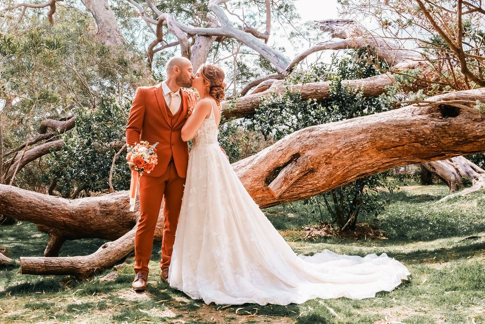 Bride and groom share a kiss in a lush garden beside a fallen tree, groom wearing a rust orange suit.