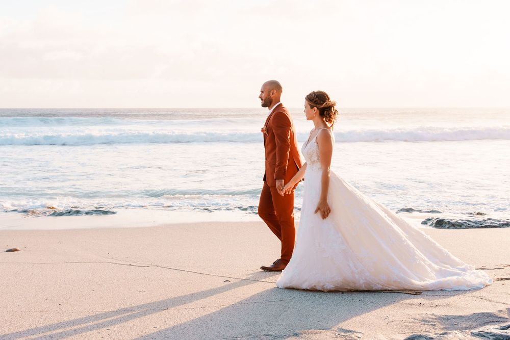 Bride and groom walk hand-in-hand on a sandy beach at sunset, waves gently crashing behind them.