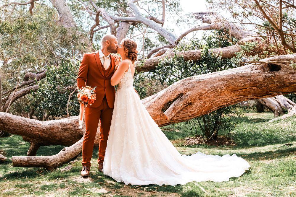 Bride and groom share a kiss outdoors near a fallen tree, groom in rust suit, bride in white lace gown with train.