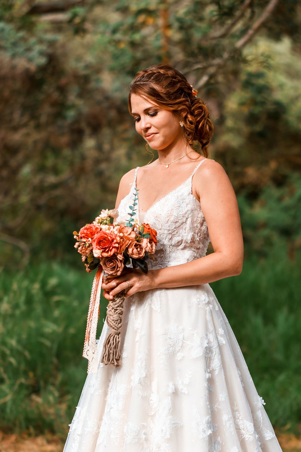 Bride in white lace gown holding orange rose bouquet outdoors, smiling softly in a garden setting.