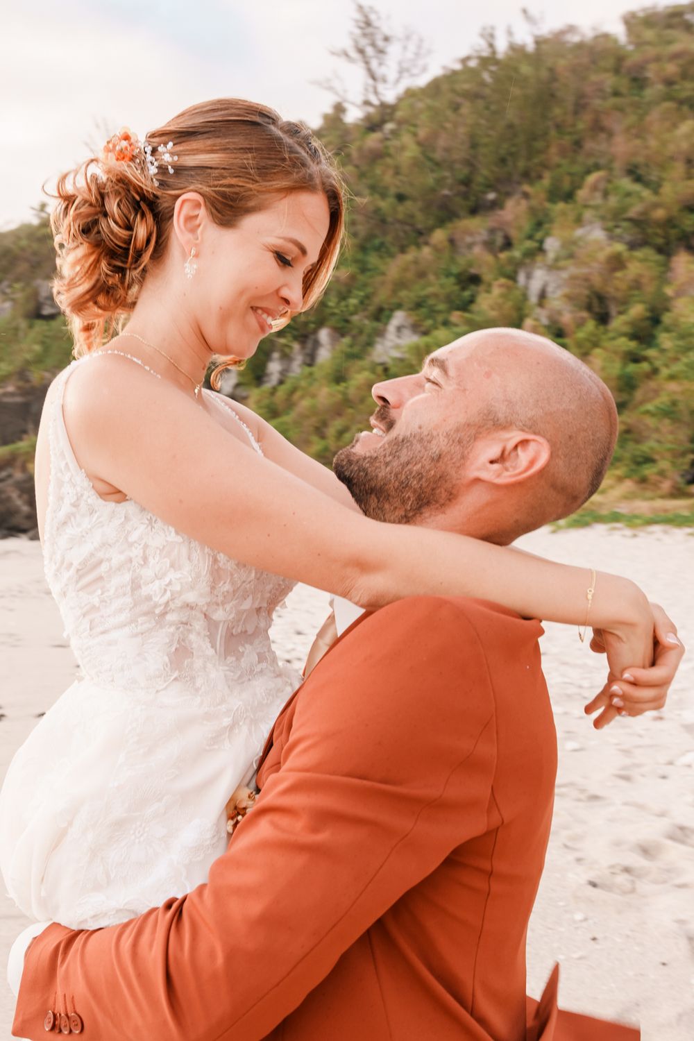 Bride and groom sharing a joyful moment on a tropical beach, groom in rust suit lifting bride in white dress.