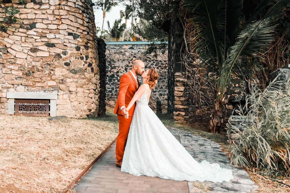 Bride in white gown and groom in orange suit share a kiss on a stone pathway surrounded by lush greenery.