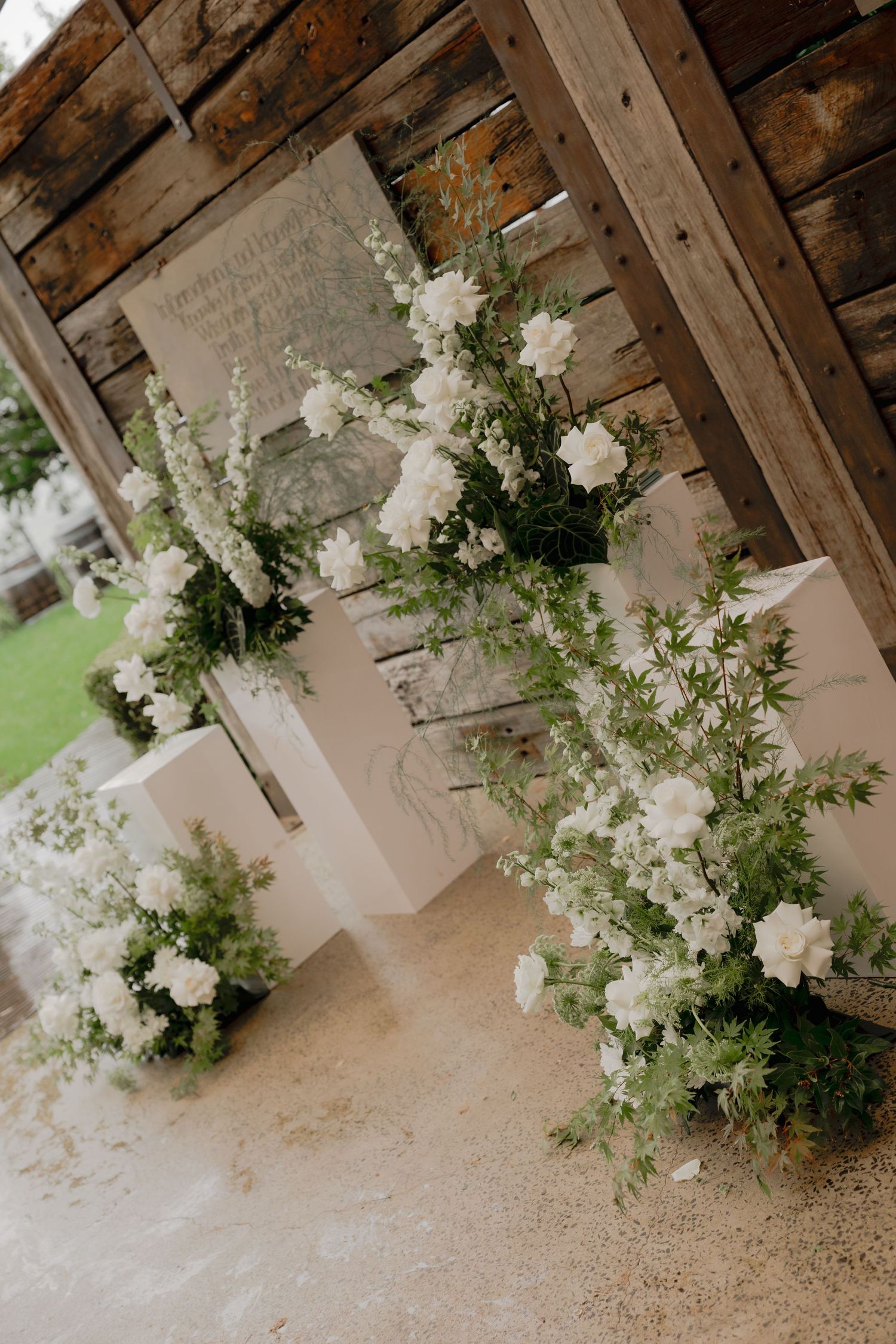 Elegant white floral arrangements with greenery on white pedestals under rustic wooden beams at a wedding venue.