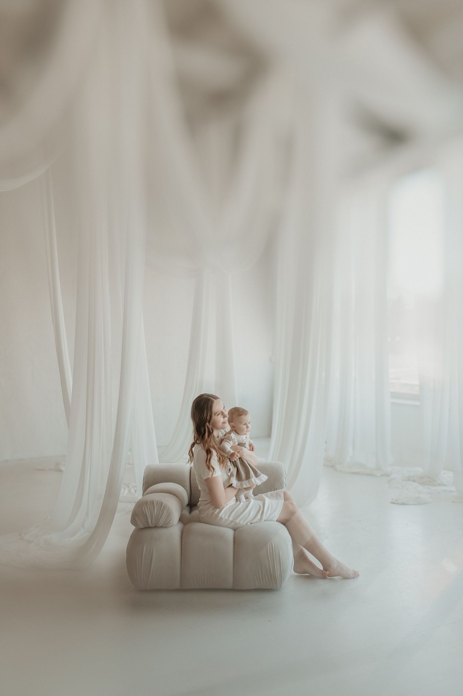 A mother sits on a white sofa holding her baby, surrounded by flowing white curtains in a bright, airy studio.