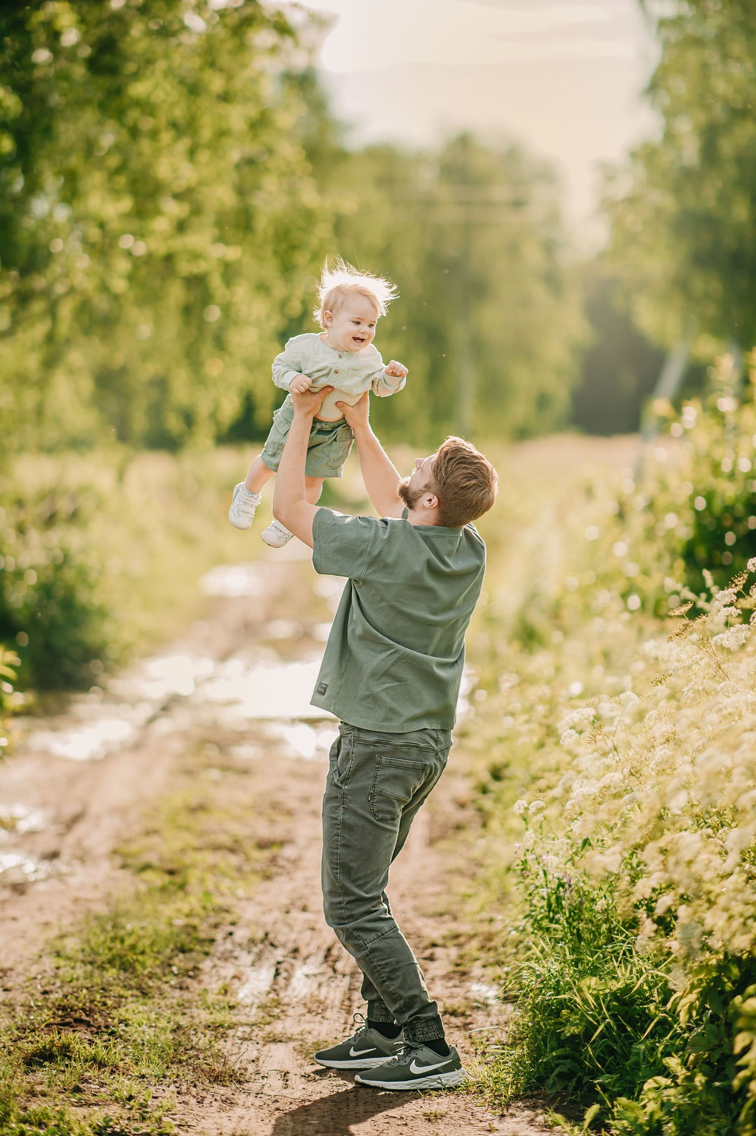 Father joyfully lifting baby in the air on a sunny rural path surrounded by lush green trees.