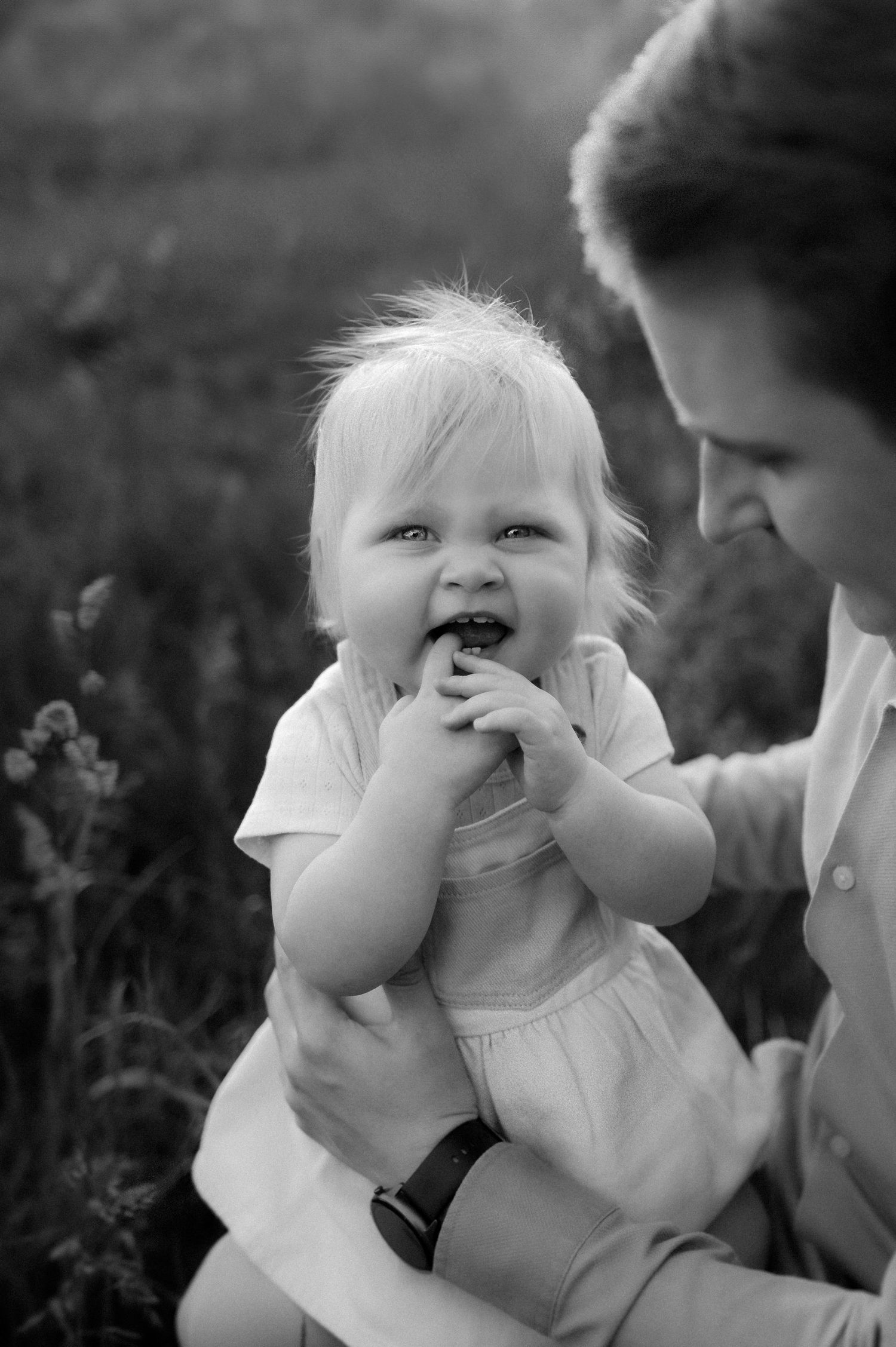Black and white photo of a smiling toddler in a dress being held by an adult outdoors in a grassy field.
