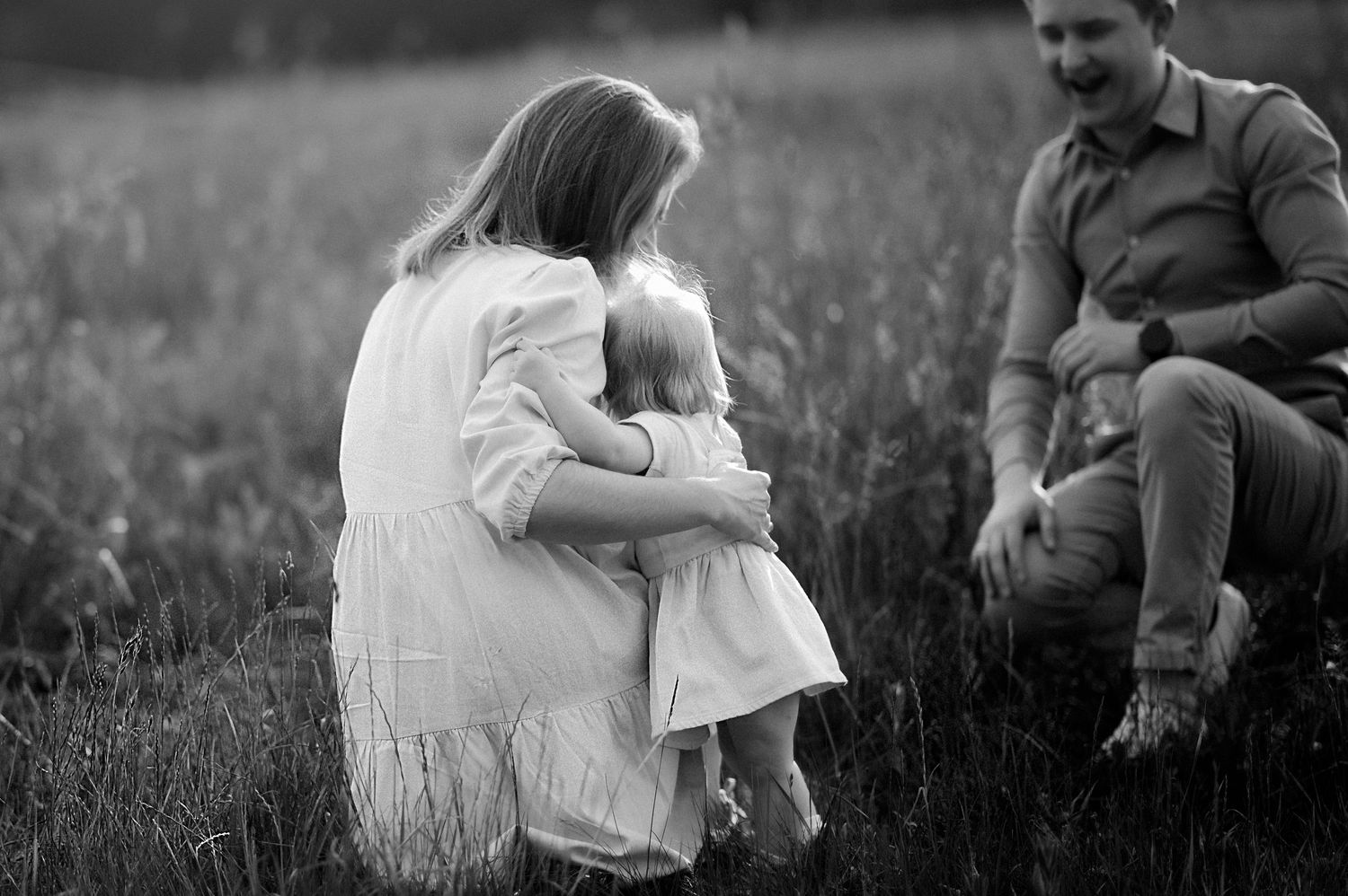 Black and white photo of a mother hugging her toddler in a field while a man sits nearby smiling.