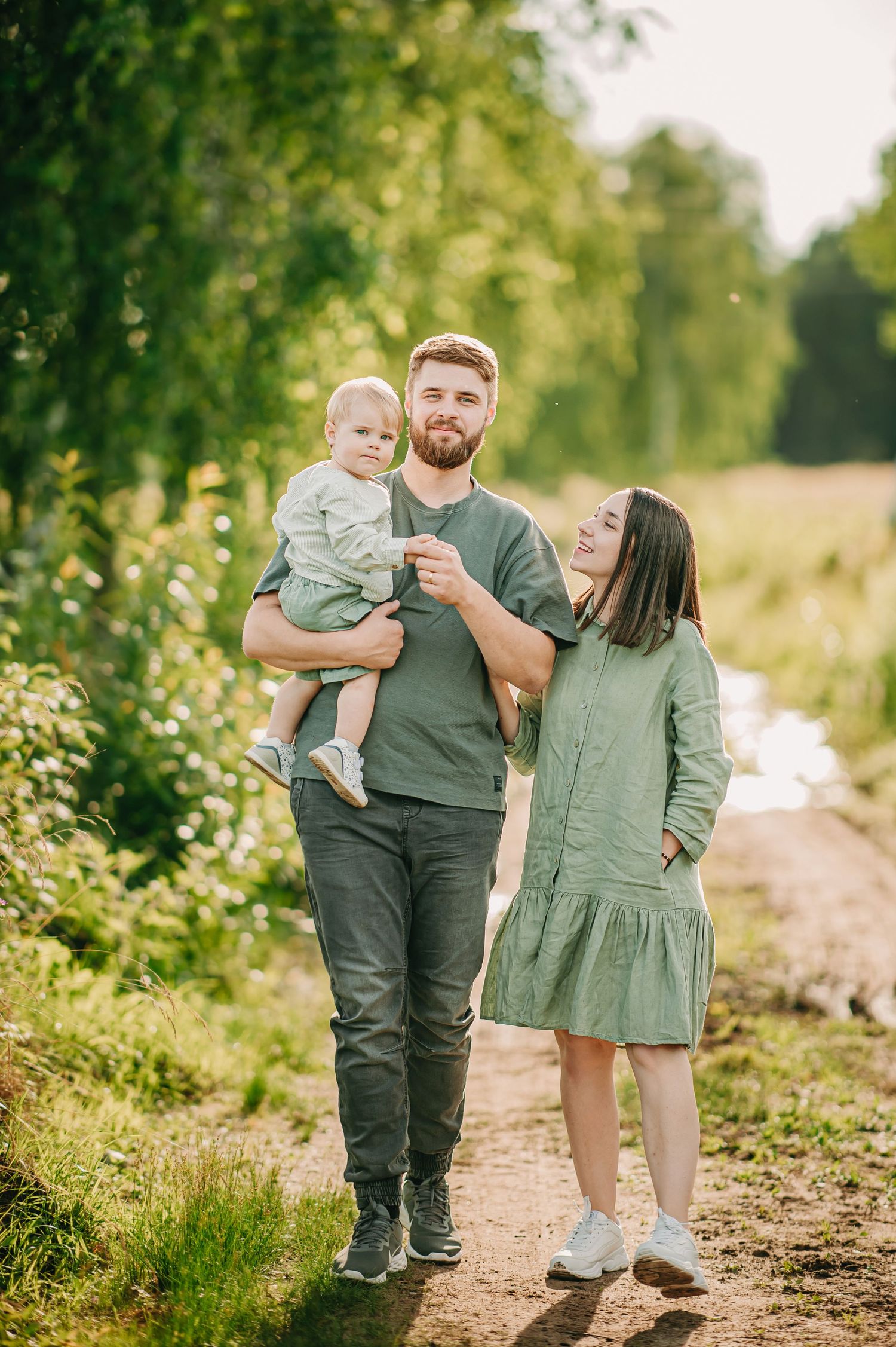 Young family in green outfits walks on a rural path, father holding baby, mother smiling beside them.