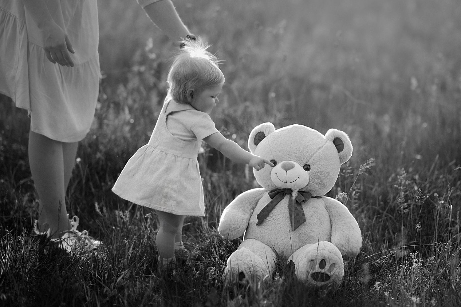 Black and white photo of a toddler girl in a dress touching a large teddy bear in a grassy field.