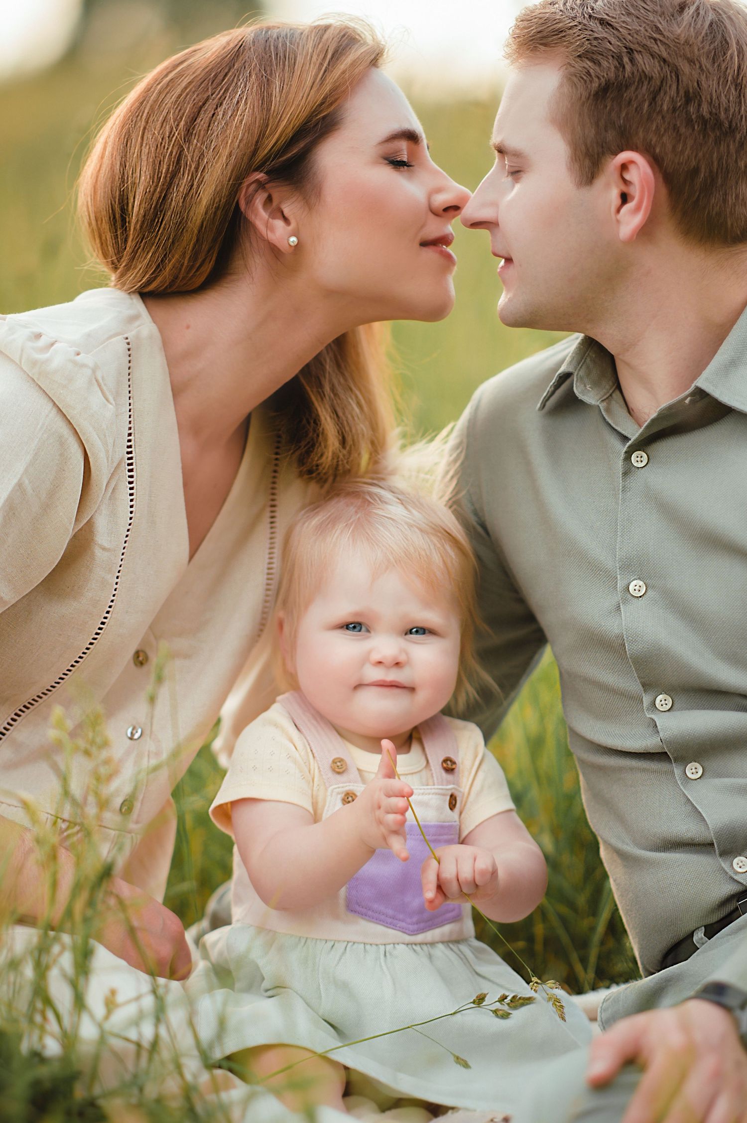 A young family sits in a green field, parents leaning in for a kiss while their toddler daughter looks at the camera.