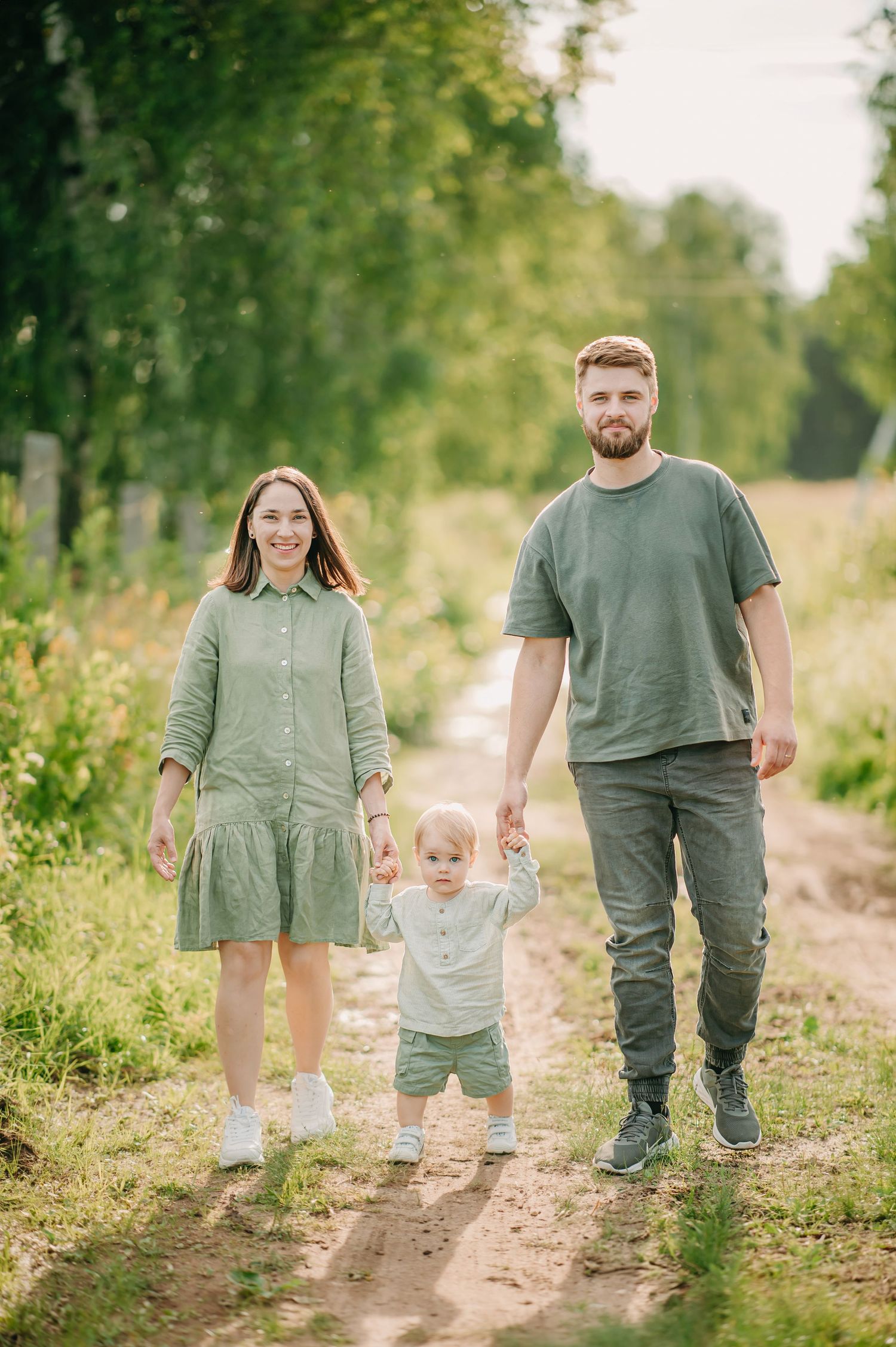 Happy young family with toddler walking on a sunny rural path surrounded by green trees and meadows.
