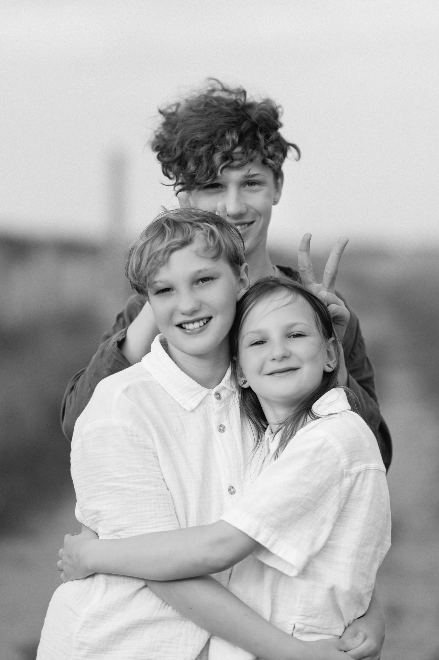 Black and white portrait of three smiling siblings hugging outdoors, oldest teen giving bunny ears.