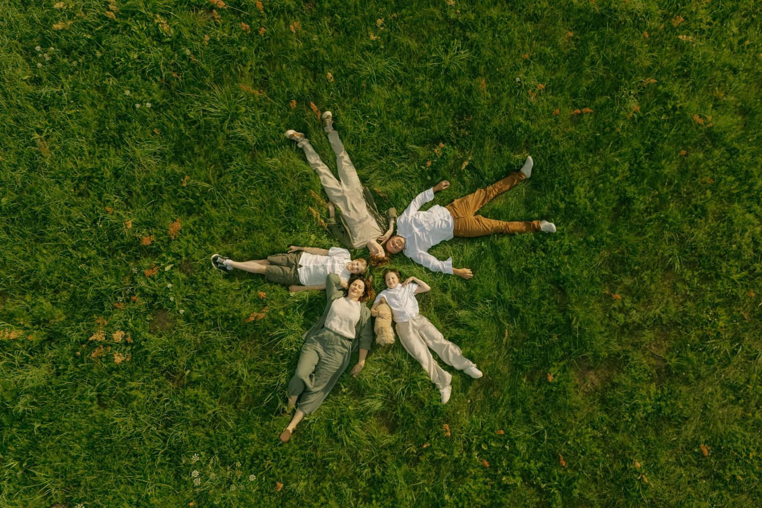Aerial view of four people lying on green grass in a star formation, wearing white and neutral outfits.
