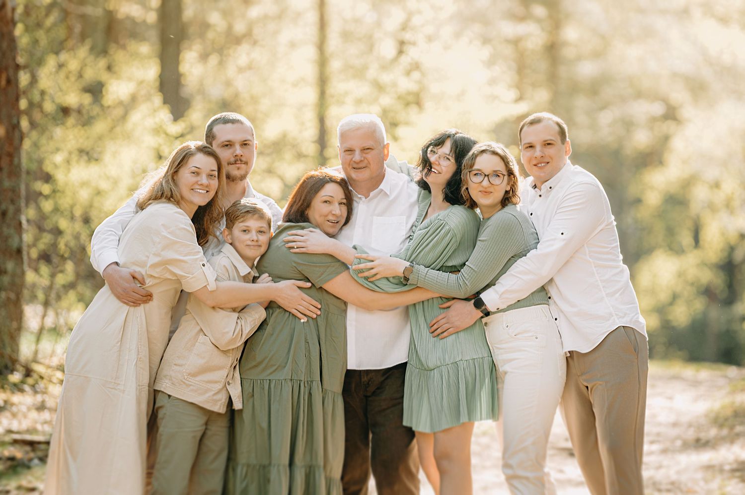 Large family group portrait outdoors in sunlit forest, adults and children embracing, wearing coordinated green and white outfits.